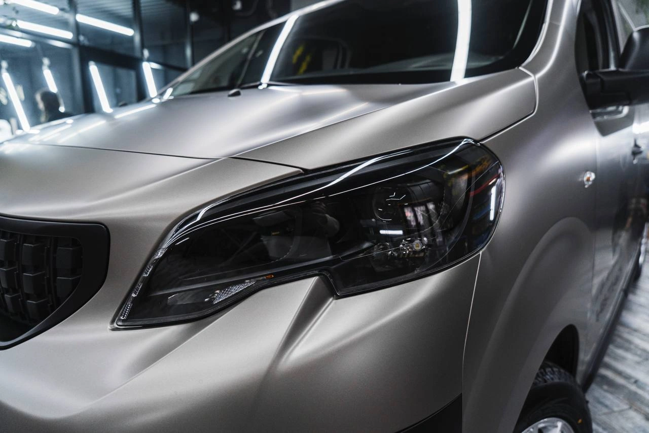 Тёмная передняя фара Peugeot Expert крупным планом на фоне серой кузовной панели с отражениями света.