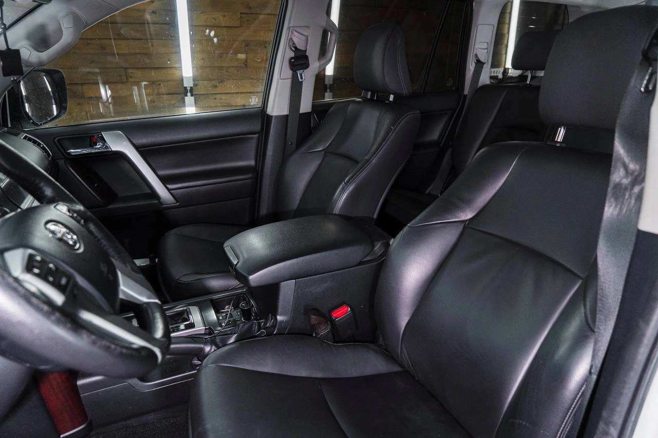 Передние сиденья Toyota Land Cruiser Prado с гладкой тёмной обивкой, рядом руль и центральный подлокотник.