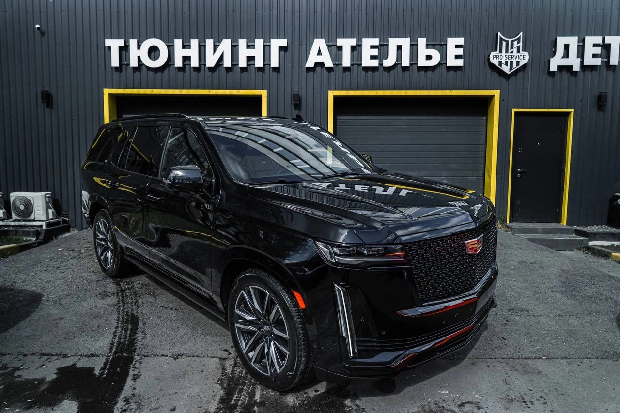 Чёрный Cadillac Escalade у входа в ателье: вид спереди и сбоку на крупную решётку и вертикальные фары.