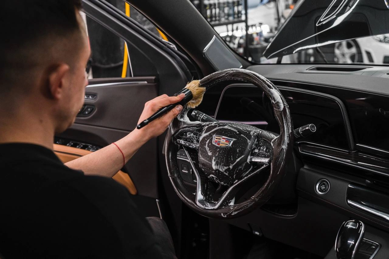 Мастер очищает рулевое колесо Cadillac Escalade небольшой щёткой внутри тёмного салона автомобиля.