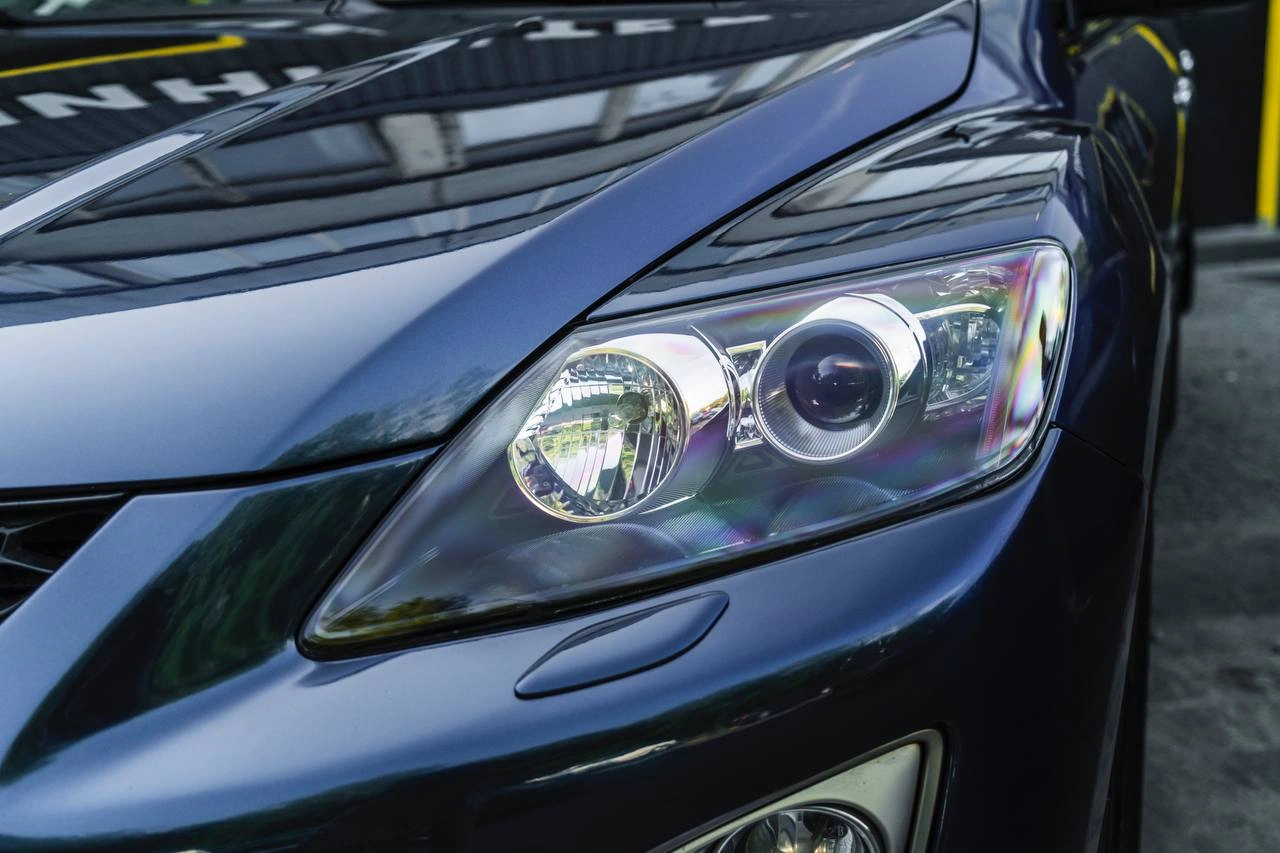 Передняя фара синей Mazda CX-7 крупным планом: прозрачное стекло, линзованный модуль и отражатели.