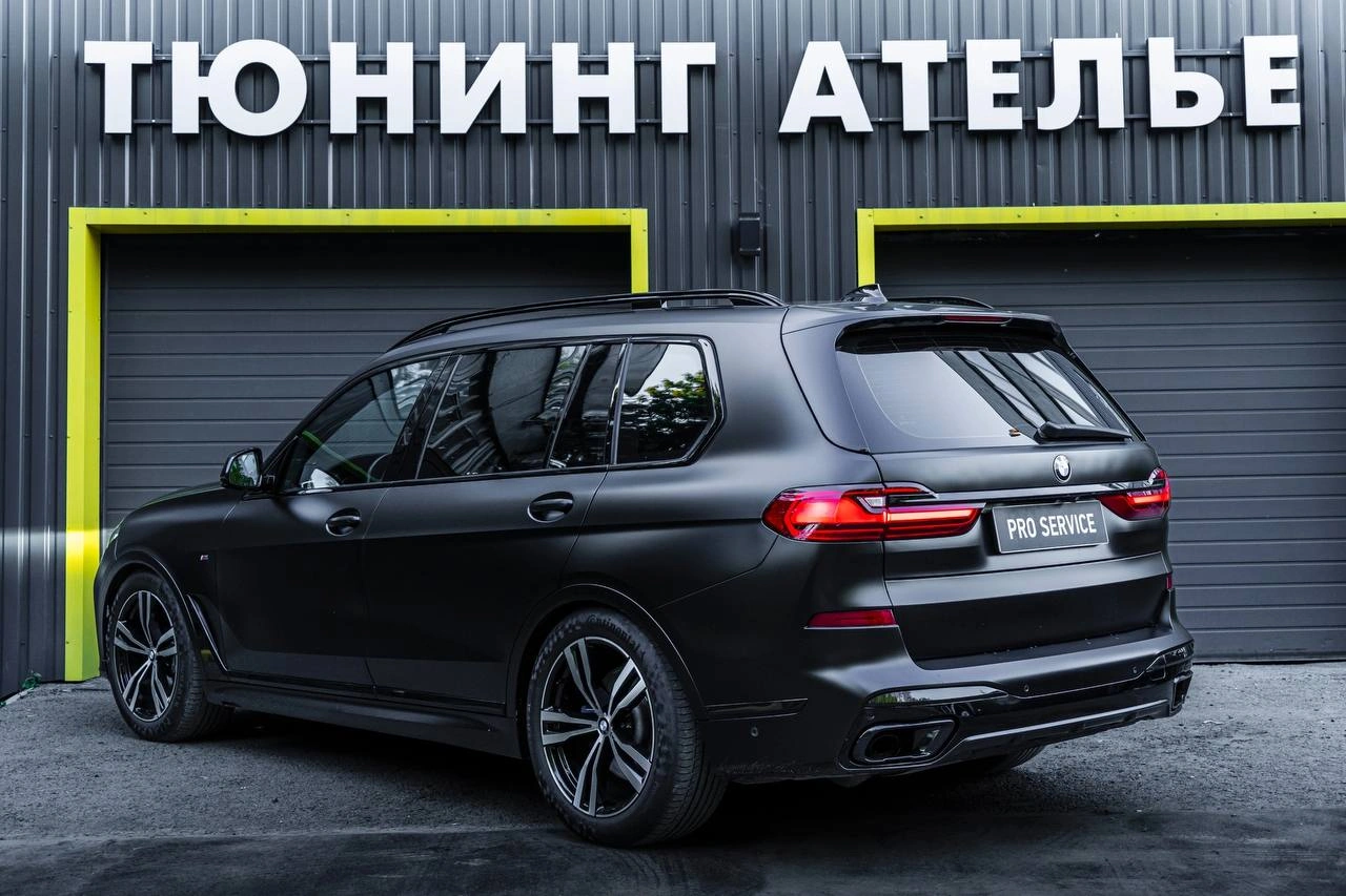 BMW X7 с тёмным кузовом, общий вид сзади под углом у ворот студии.