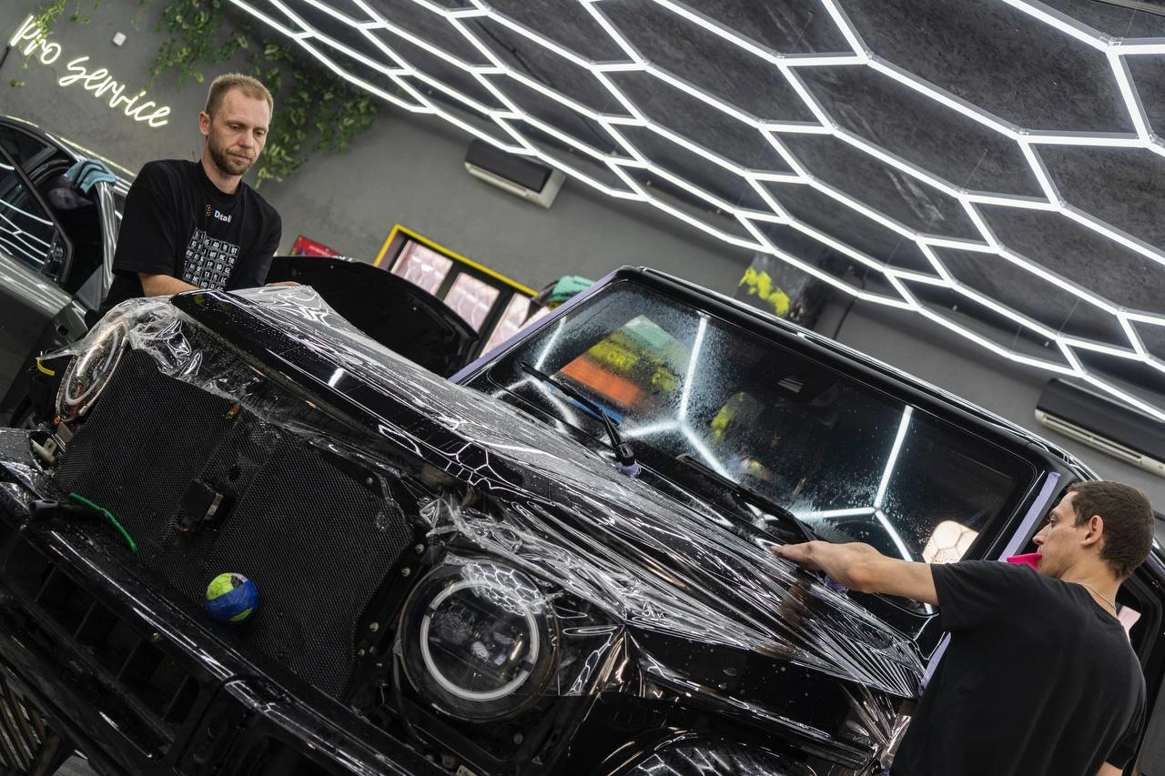 Два мастера натягивают прозрачную плёнку на капот Mercedes-benz G63 AMG в рабочем боксе студии.