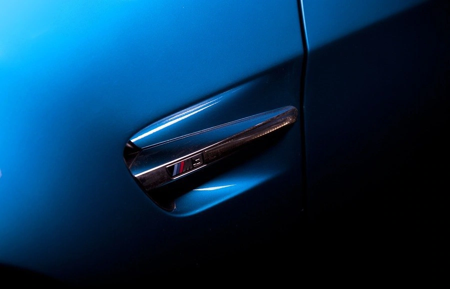 Боковая кузовная деталь синего BMW M3 крупным планом: блестящая вставка с эмблемой M3 и дверной зазор.