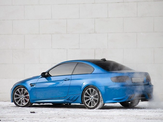 Синий BMW M3 на заснеженной площадке у белой стены: вид со стороны заднего крыла и боковых окон.
