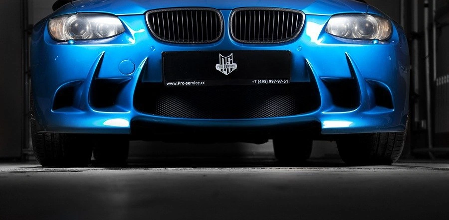 Низкий ракурс спереди на BMW M3: синяя передняя часть, тёмные воздухозаборники и фары в освещённом боксе.