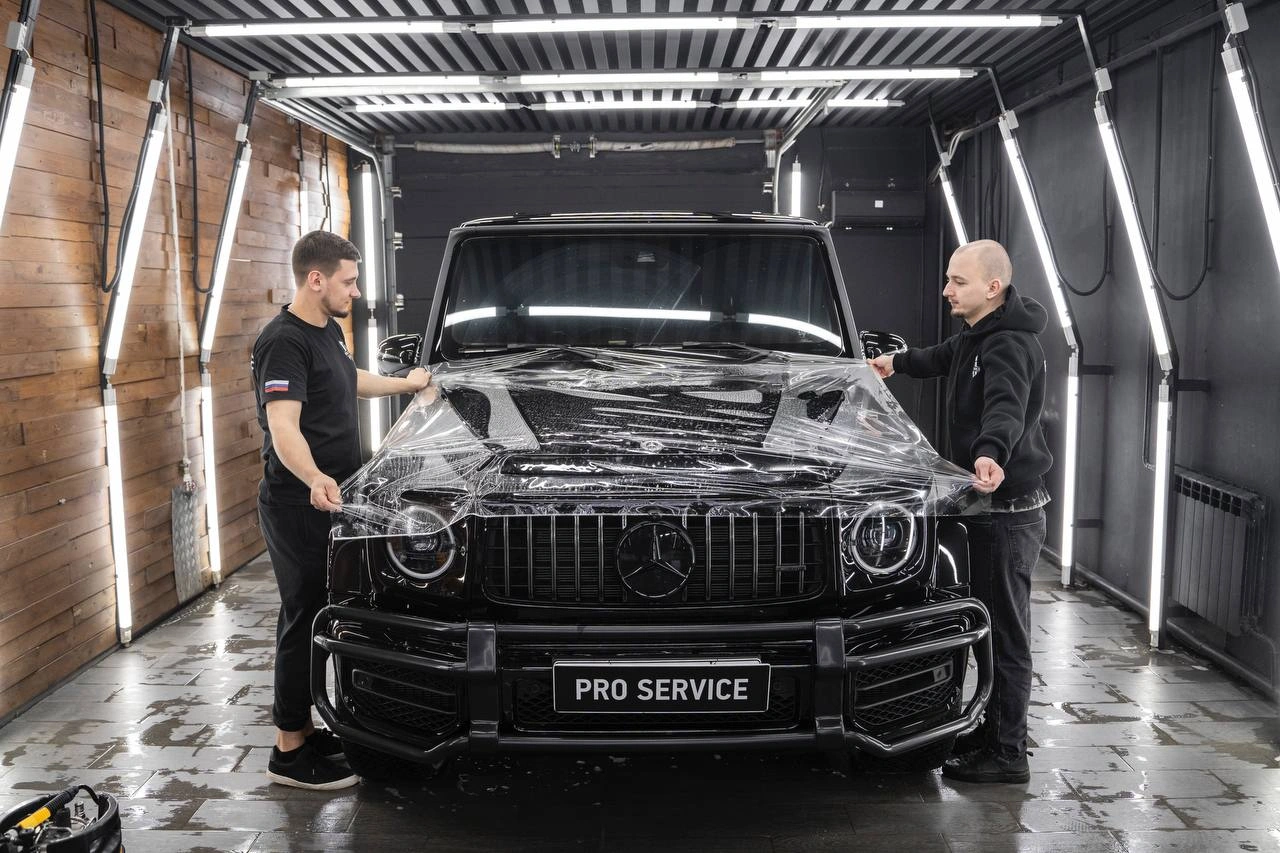 Двое мастеров натягивают прозрачную плёнку на капот чёрного Mercedes-Benz G-class.