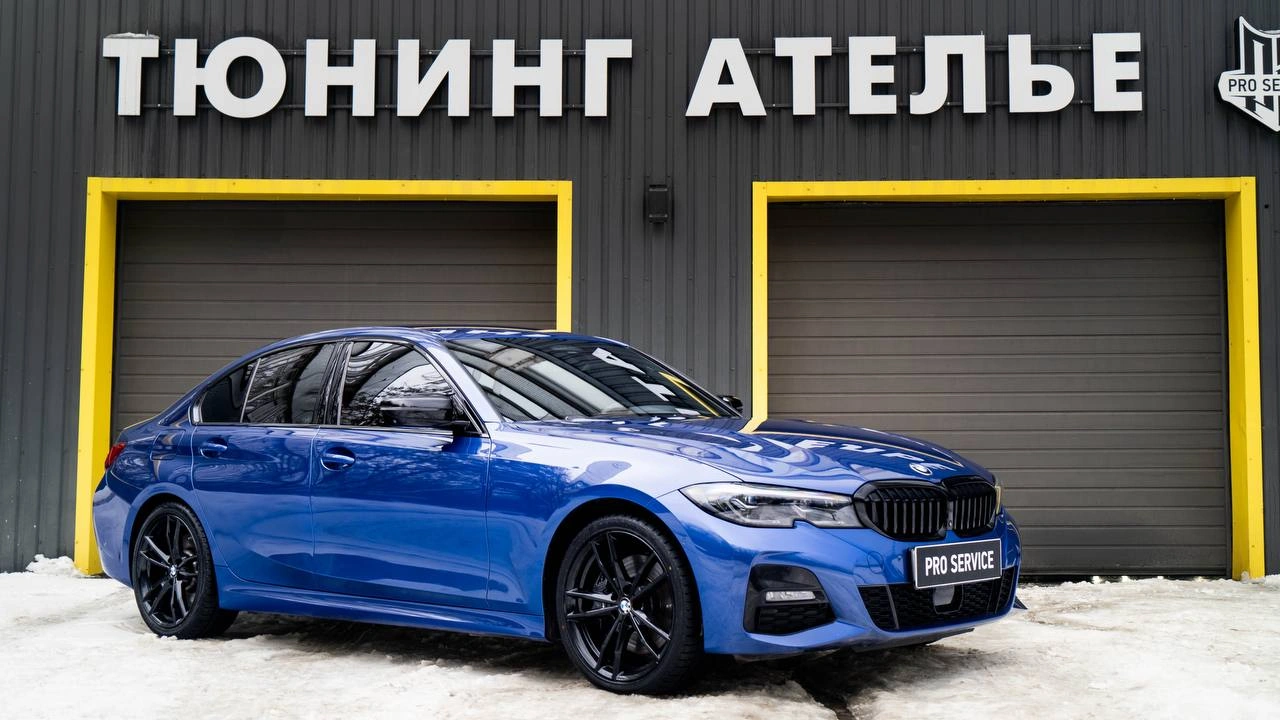 Синий BMW 3-й серии с чёрными колёсами на снегу у входа в студию, вид спереди под углом.