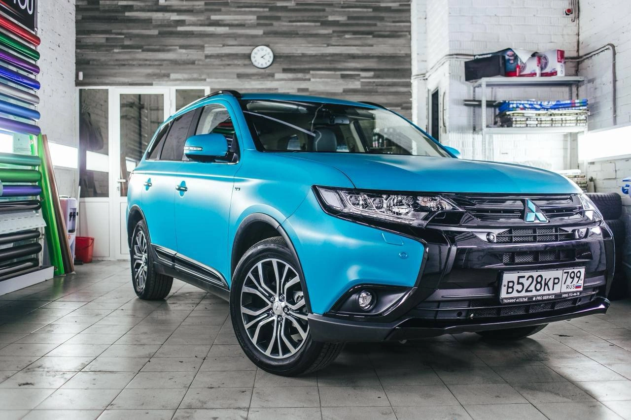 Голубой Mitsubishi Outlander в рабочем помещении: вид спереди и сбоку с чёрной передней частью и светлыми дисками.