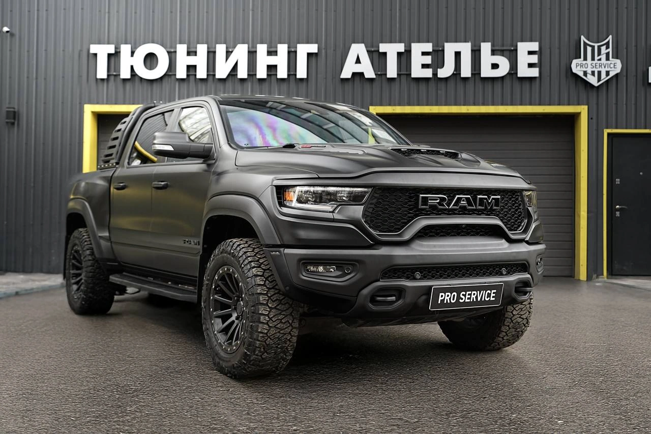 Серый Dodge Ram TRX у ворот ателье: вид спереди и сбоку с крупной решёткой RAM и внедорожными шинами.