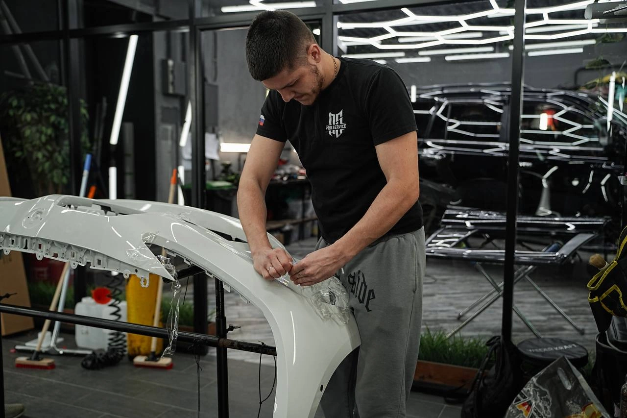 Мастер подгоняет плёнку на снятом белом бампере BMW М4, установленном на рабочей стойке.