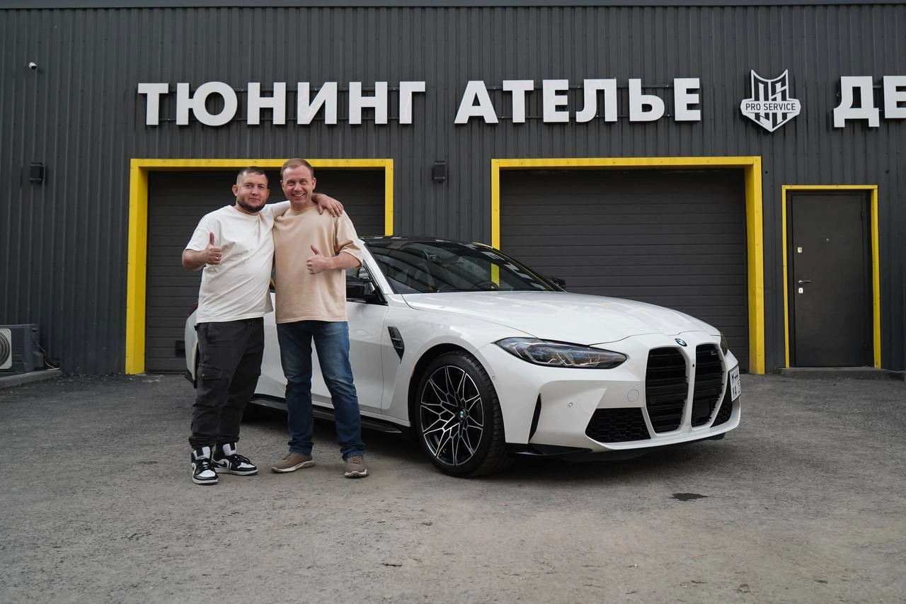 Белая BMW М4 у входа в студию; рядом с автомобилем стоят двое мужчин.