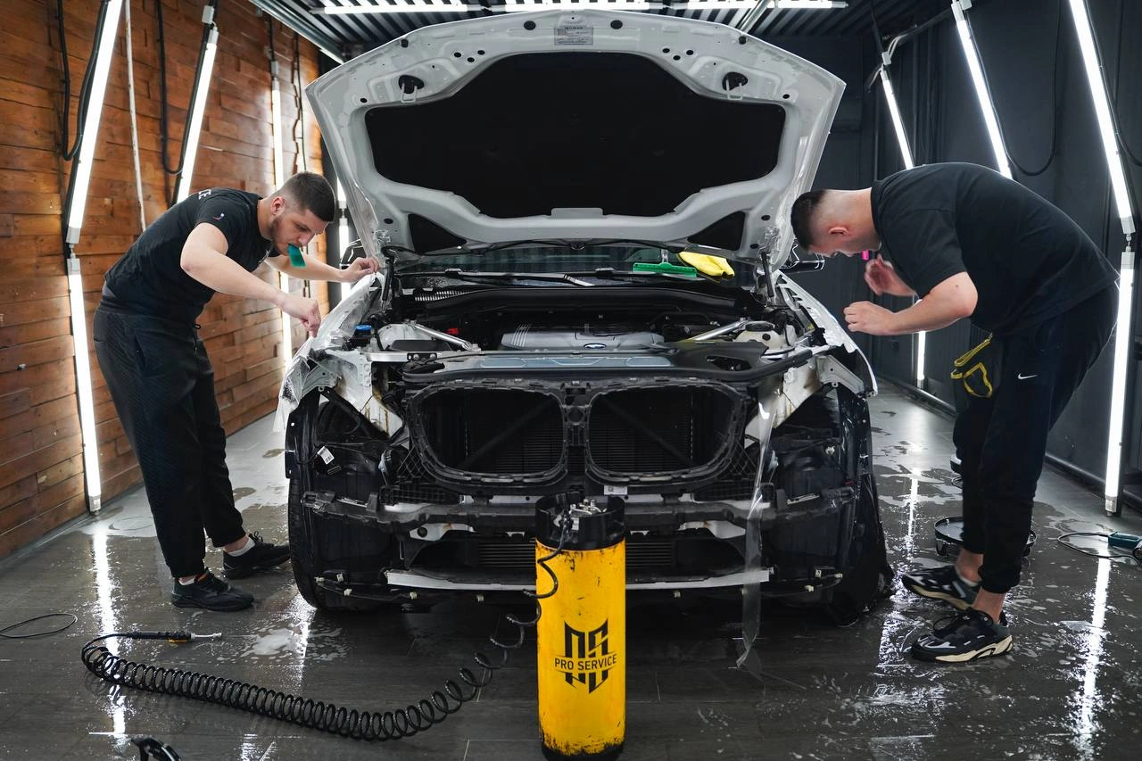 Два мастера очищают переднюю часть BMW X3: капот открыт, передний бампер демонтирован.