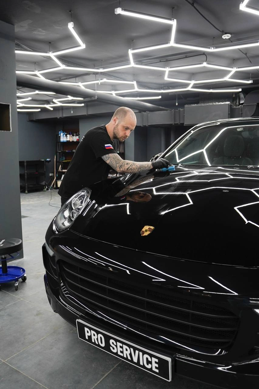 Мастер работает голубым аппликатором на капоте Porsche Cayenne у основания лобового стекла.