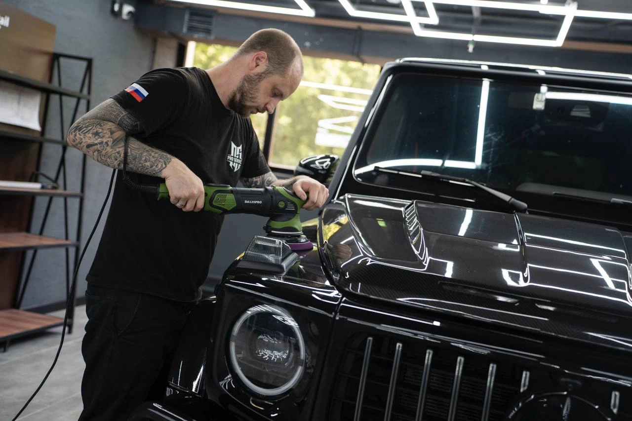 Мастер полирует капот Mercedes-Benz G-class AMG рядом с круглой фарой и решёткой чёрного внедорожника.