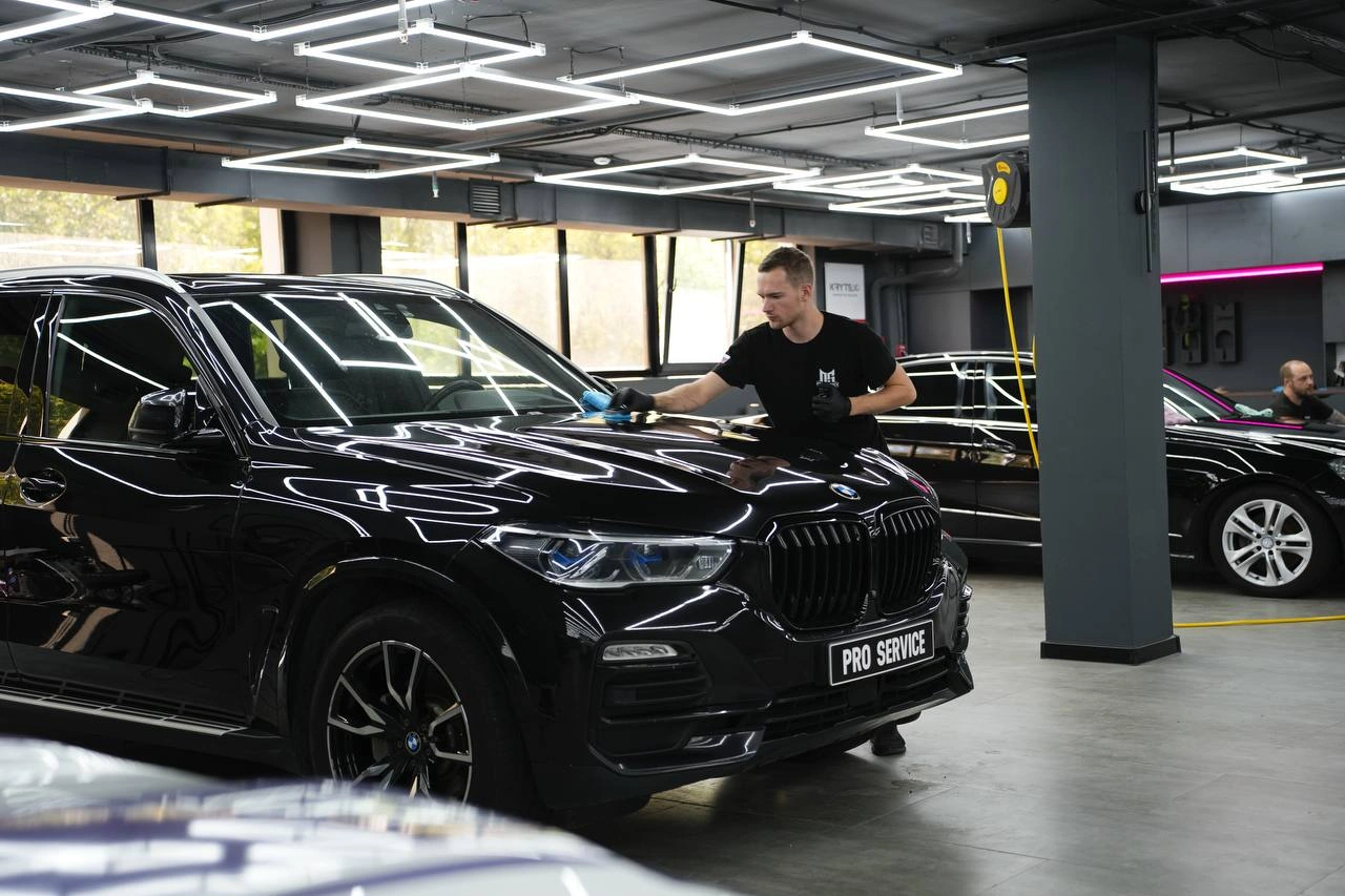 Мастер обрабатывает капот чёрного BMW X5; на кузове и лобовом стекле отражаются потолочные световые линии.