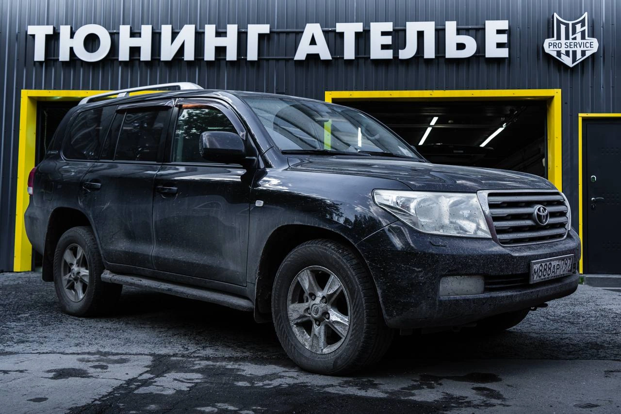 Toyota Land Cruiser 200 у ворот ателье: вид спереди и сбоку с серебристой решёткой радиатора.