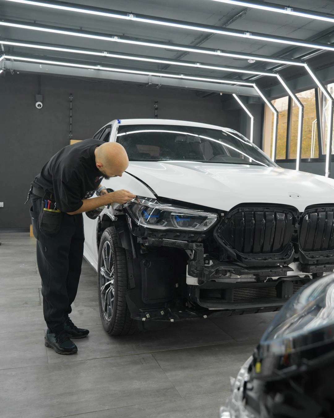 Мастер подгоняет плёнку у передней фары белого BMW X6; бампер снят для доступа к кузовным кромкам.