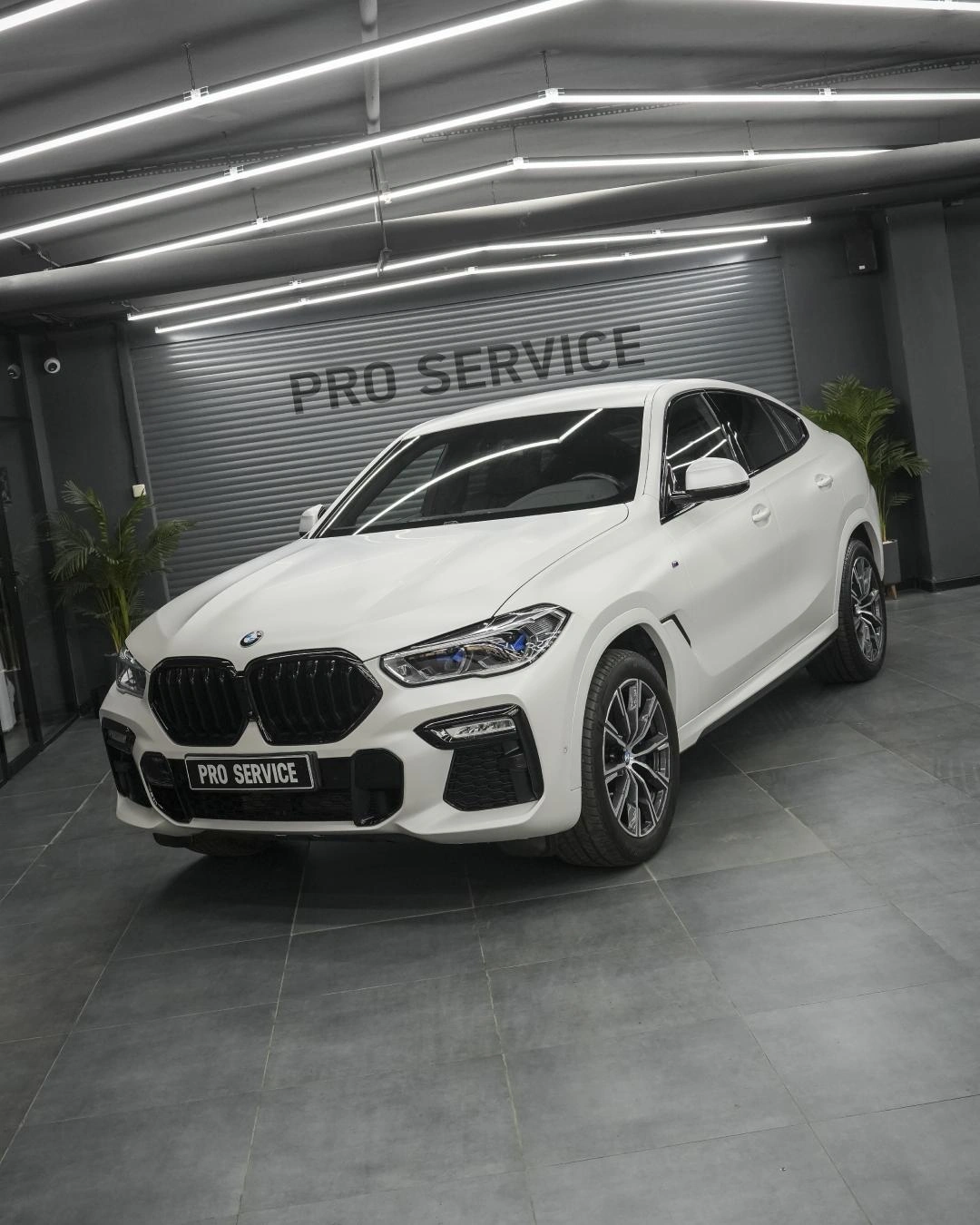 Белый BMW X6 в боксе PRO Service: вид спереди и сбоку с тёмной решёткой радиатора и светлыми колёсами.