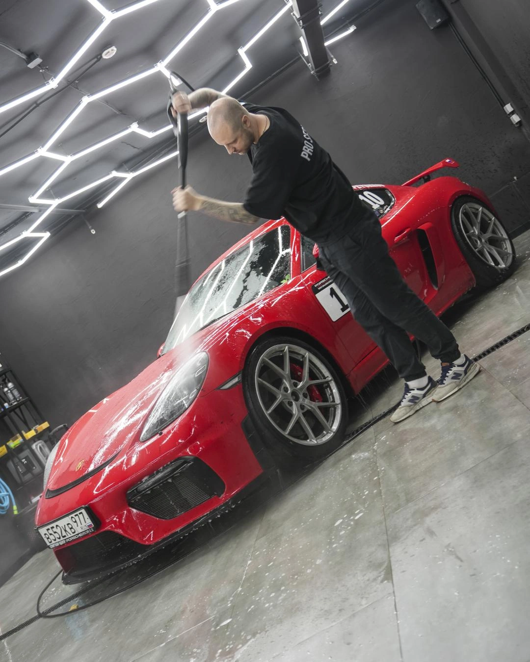 Мастер моет кузов Porsche Cayman GT-4 струёй воды; красный автомобиль находится в боксе студии.