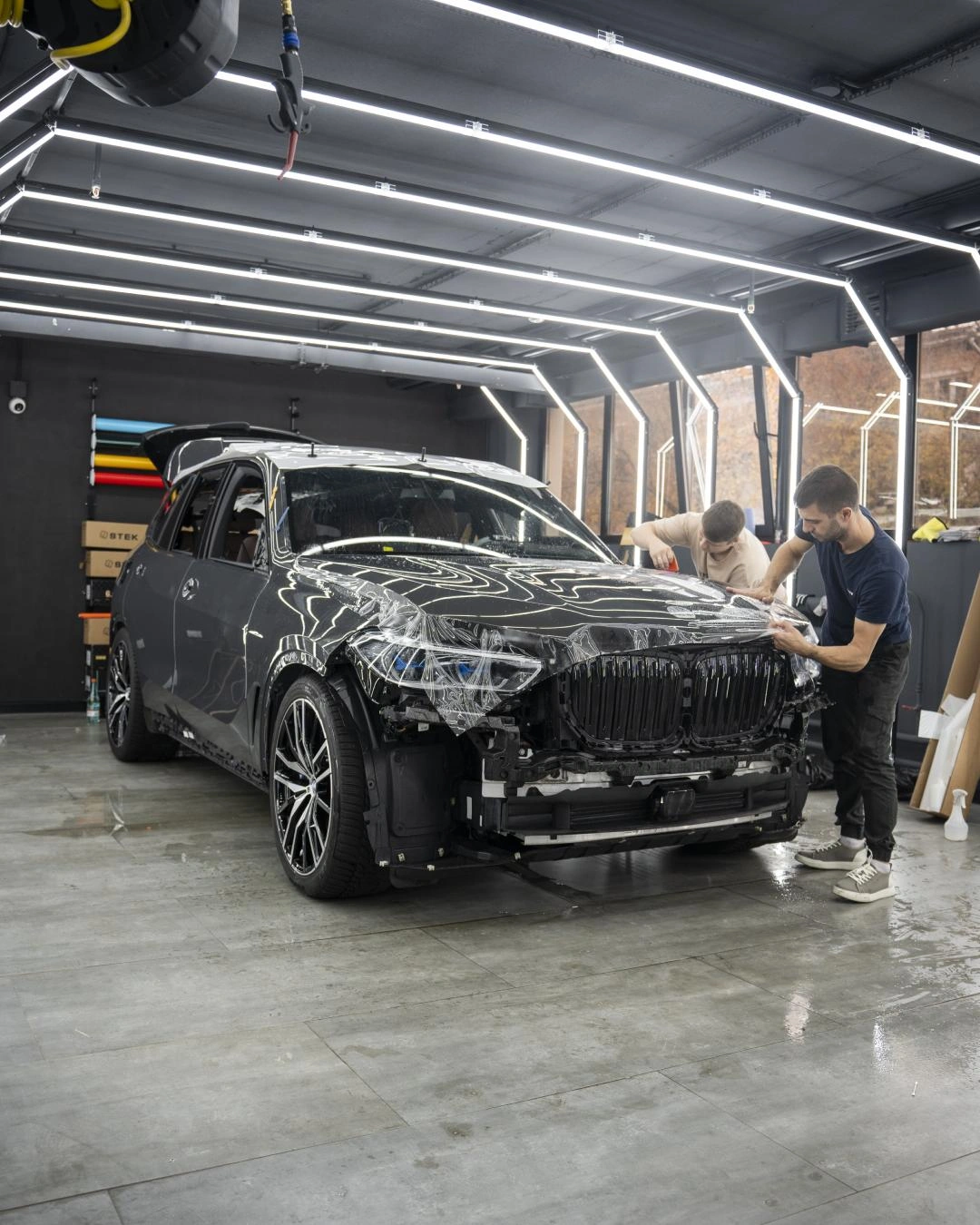 Мастера оклеивают переднюю часть BMW X5: капот покрыт прозрачной плёнкой, бампер демонтирован.