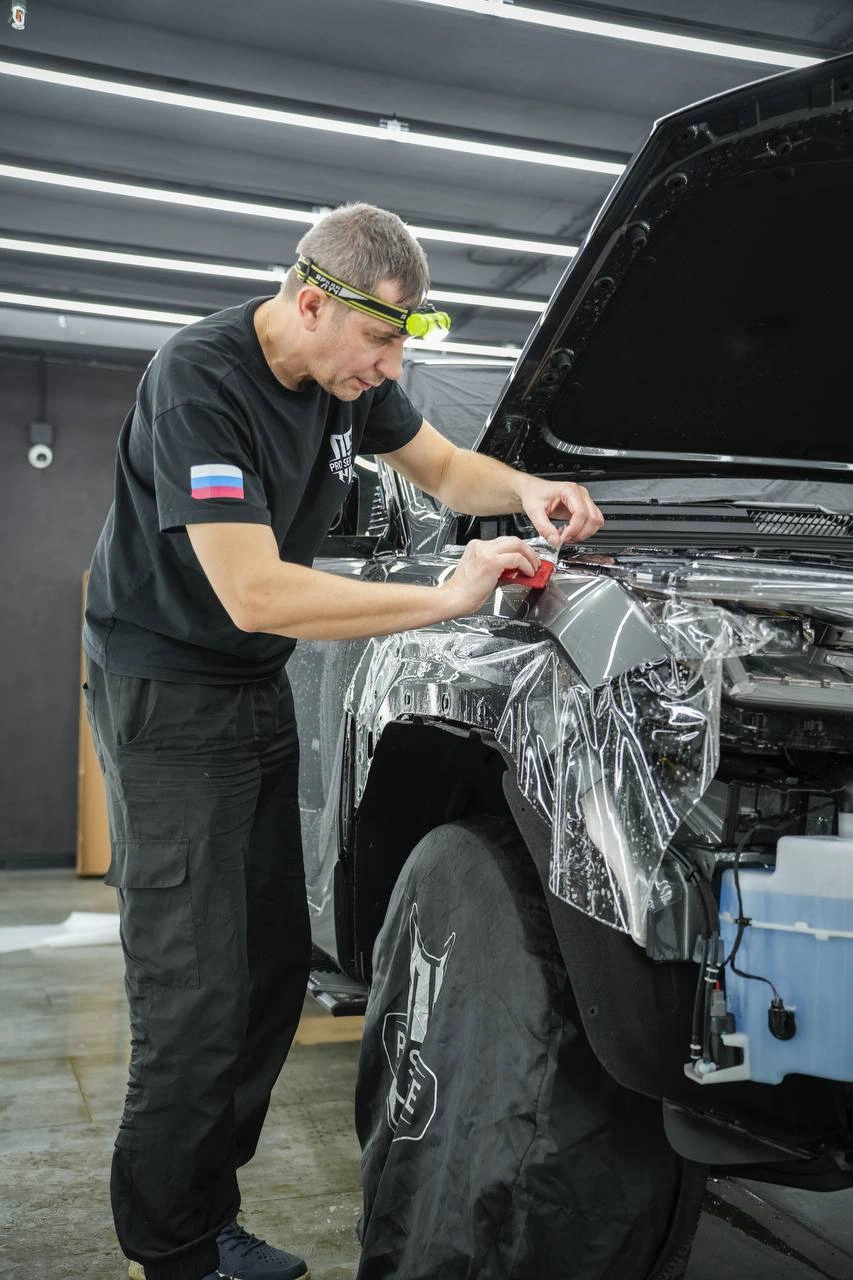 Мастер подгоняет прозрачную плёнку на переднем крыле Tank 400; капот открыт, передняя часть разобрана.