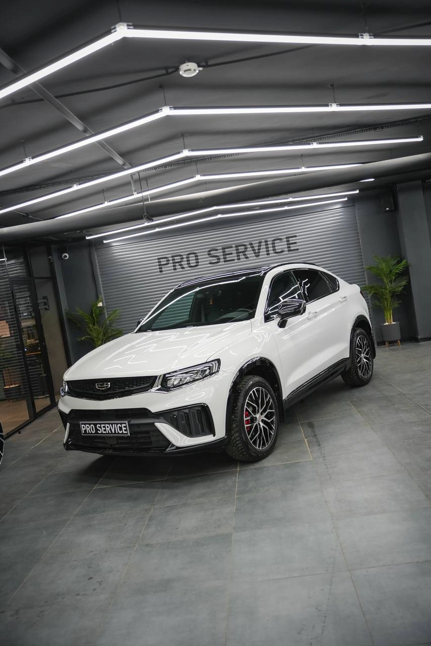 Белая Geely Tugella, общий вид спереди под углом на фоне ворот студии.