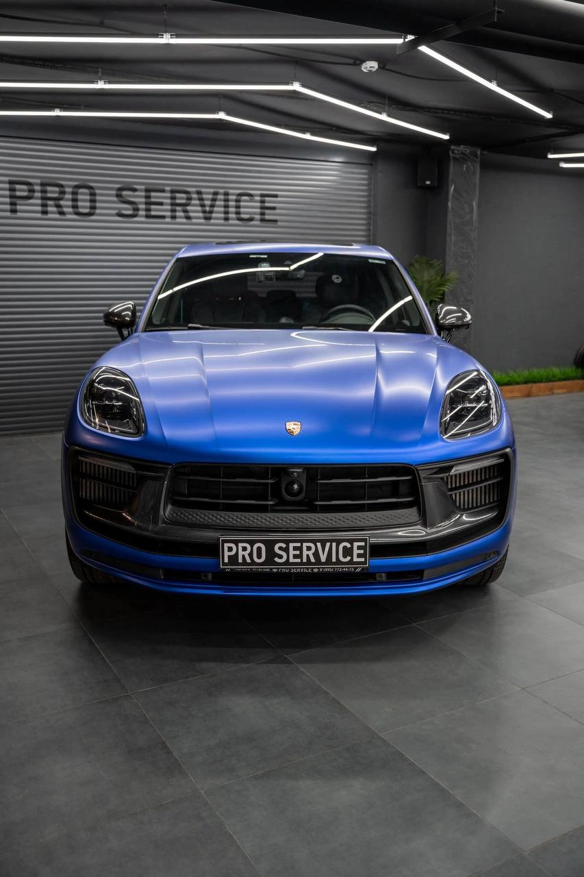 Синий Porsche Macan, прямой вид спереди с отражениями потолочного освещения на капоте.
