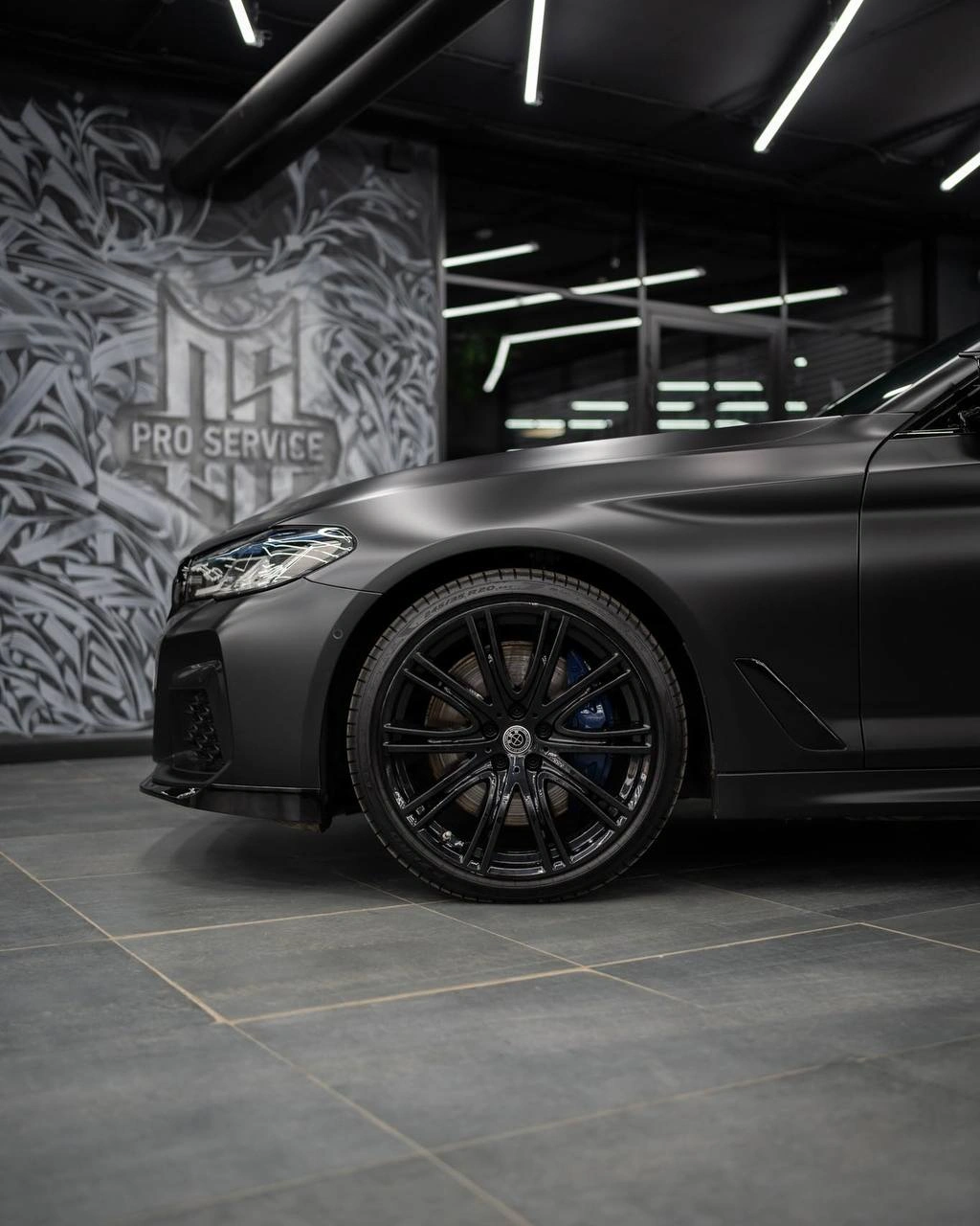 Переднее крыло и чёрное колесо BMW 5 G30 в студии: на тёмной панели отражаются потолочные светильники.