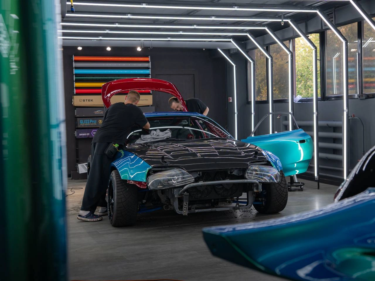 Мастер у Toyota Supra со снятым передним бампером во время оклейки: бирюзовая плёнка на кузове.