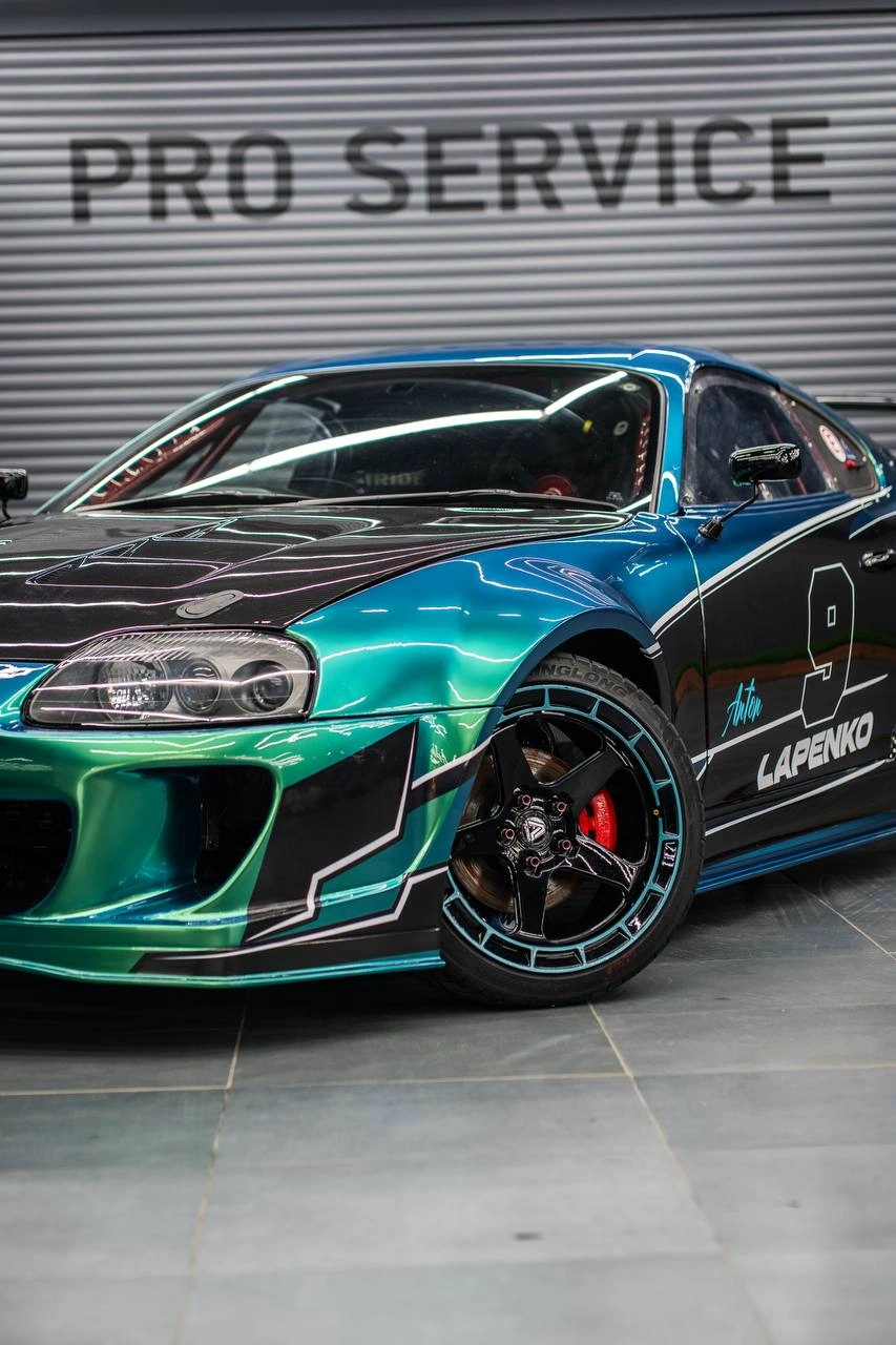 Toyota Supra с бирюзово-зелёной графикой: передний бампер, фара и чёрное колесо крупным планом.
