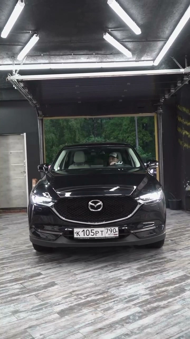 Кадр из видео: MAZDA CX-5 — тёмный кроссовер спереди в рабочем боксе под потолочным освещением
